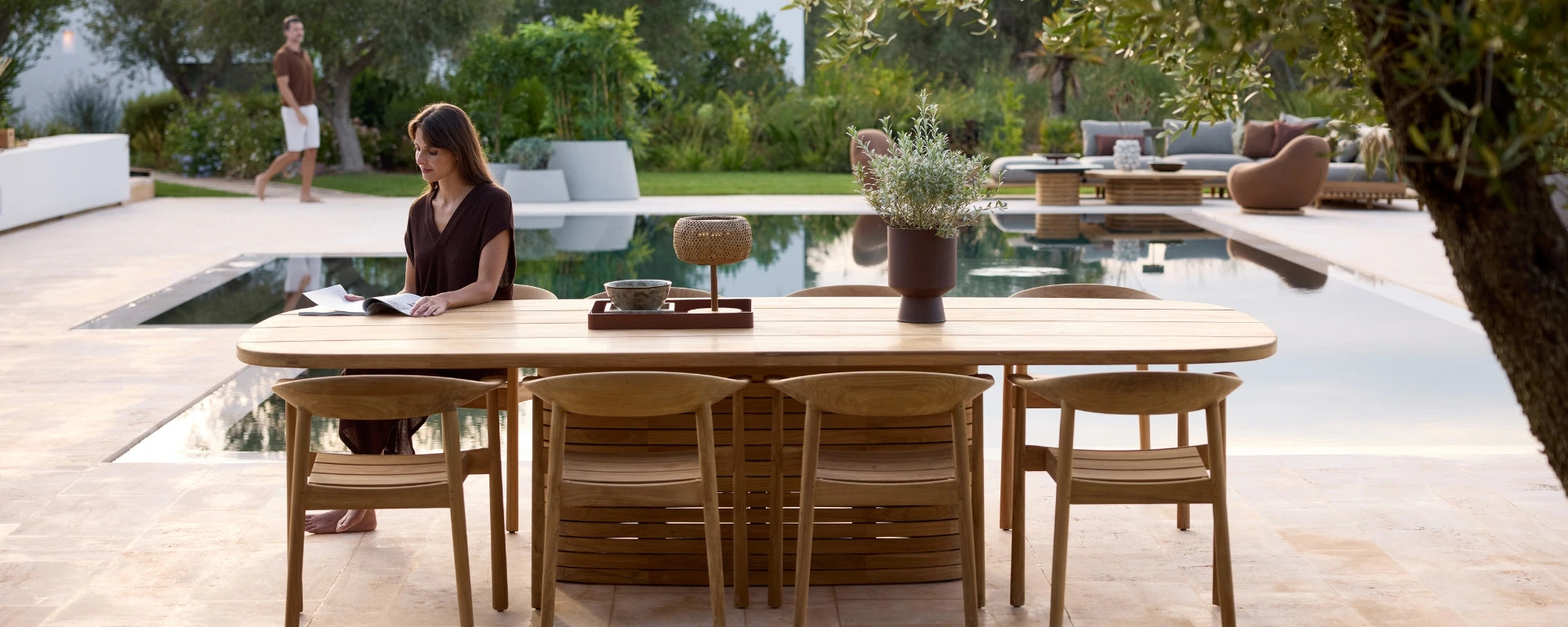 Lady sitting at oval outdoor wooden dining table with curved teak chairs next to pool