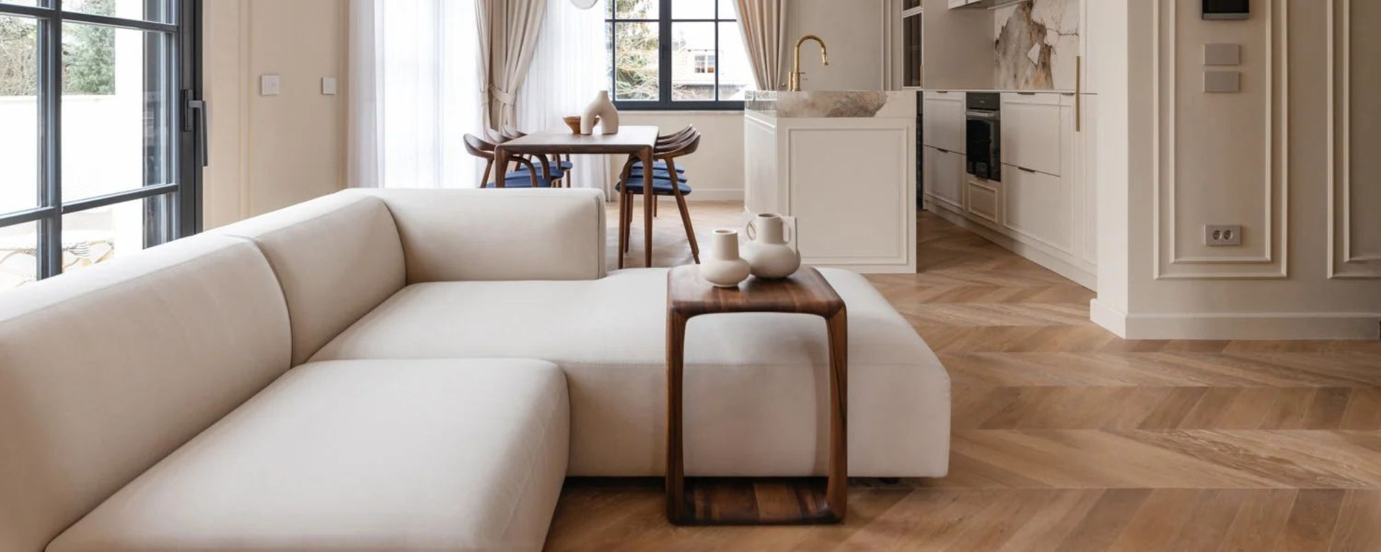 Modern living room with a white sectional sofa, wooden coffee table, and kitchen in the background.