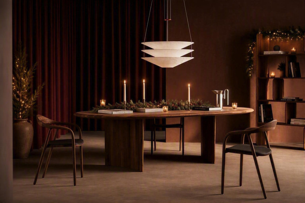 A modern dining room scene featuring a round wooden dining table with a thick pedestal base, surrounded by two dark wood chairs with black seats. A tiered white pendant light hangs above, and the table is decorated with candles and greenery.