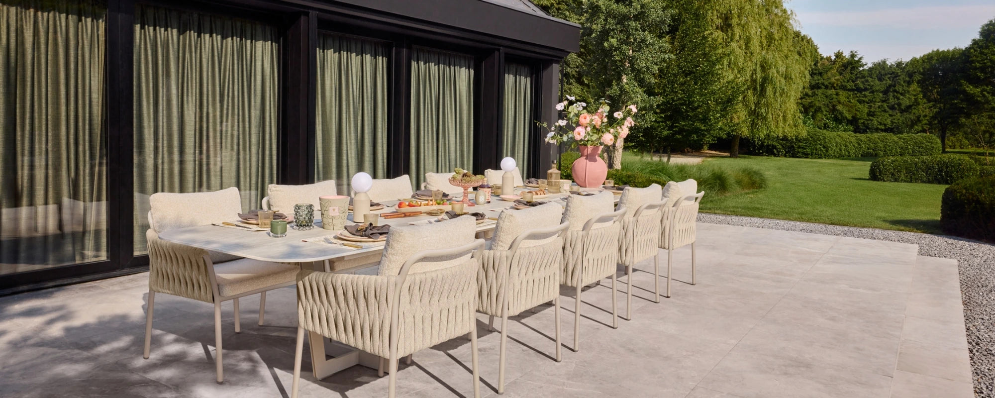 Outdoor dining setup with a long low outdoor table and chairs on a patio, surrounded by greenery.