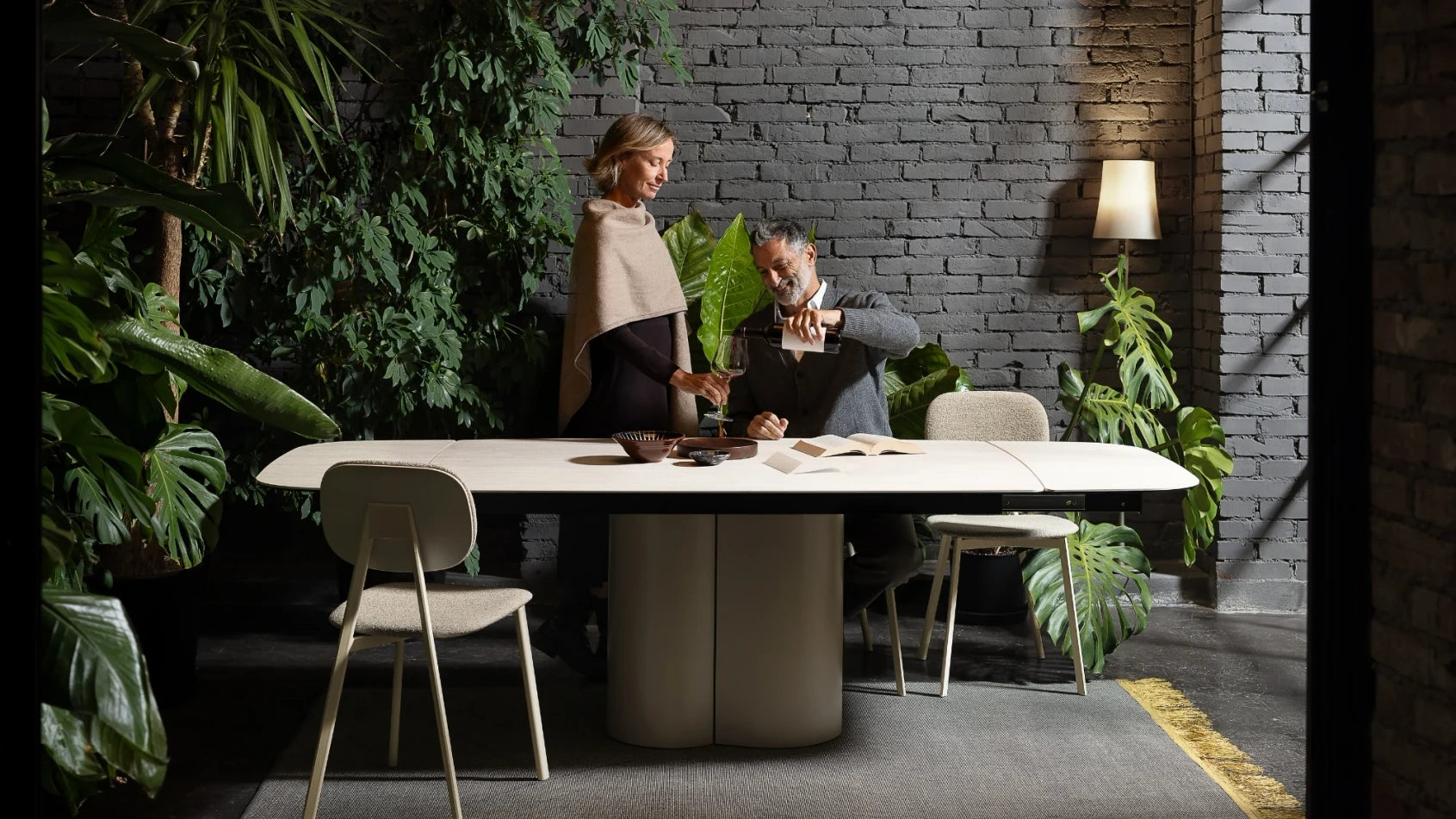 A modern dining table set in a stylish interior with a dark brick wall, lush green plants, and two people standing behind the table.