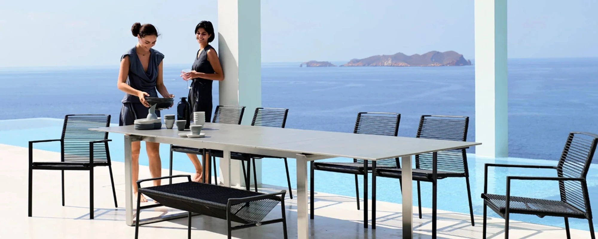 Two women standing by a large outdoor dining table with chairs on a patio overlooking the ocean.