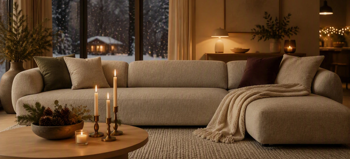A large beige sectional sofa in a cozy, warmly lit living room with a window showing a snowy outdoor view. The sofa is styled with neutral and dark accent pillows and a cream throw blanket draped over the chaise.